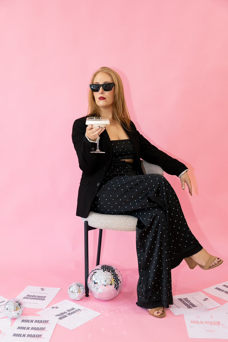 a photo of a caucasian woman sitting in a white chair in front of a pink backdrop holding a glass of milk in hand with sunglasses on looking to the side of the camera