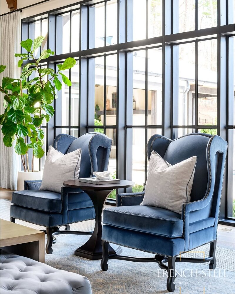 Two blue velvet armchairs with white pillows beside a round table, set near tall industrial-style windows and a fiddle leaf fig.
