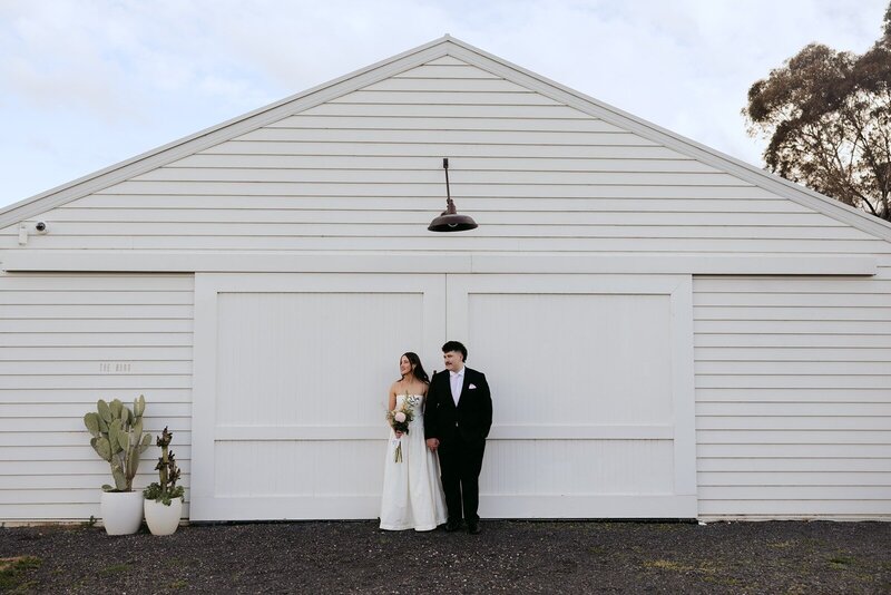 A bride and groom standing in front of a white weatherboard barn