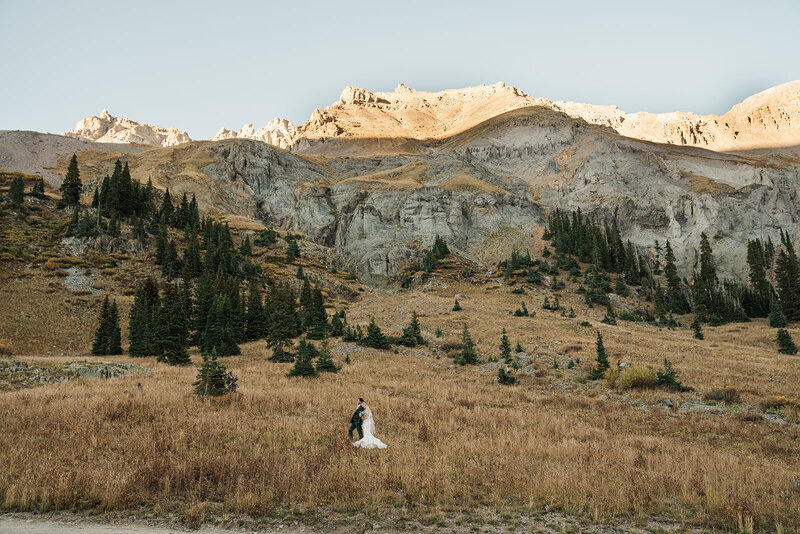A man holds a woman in a wedding dress in front of a lake and mountains