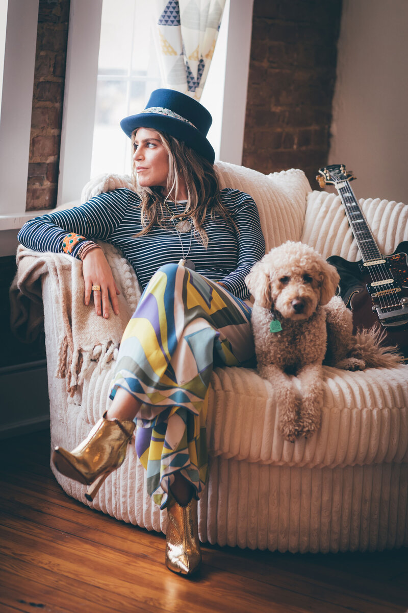 A woman in a hat sitting on a chair next to a guitar, captured by a skilled personal branding photographer Britt Elizabeth.