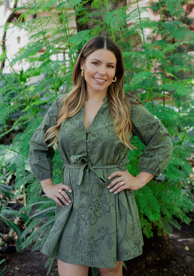 Natalie Bernachi, a smiling woman standing confidently in front of lush greenery, wearing a belted olive green eyelet dress with gold jewelry and soft waves in her hair.
