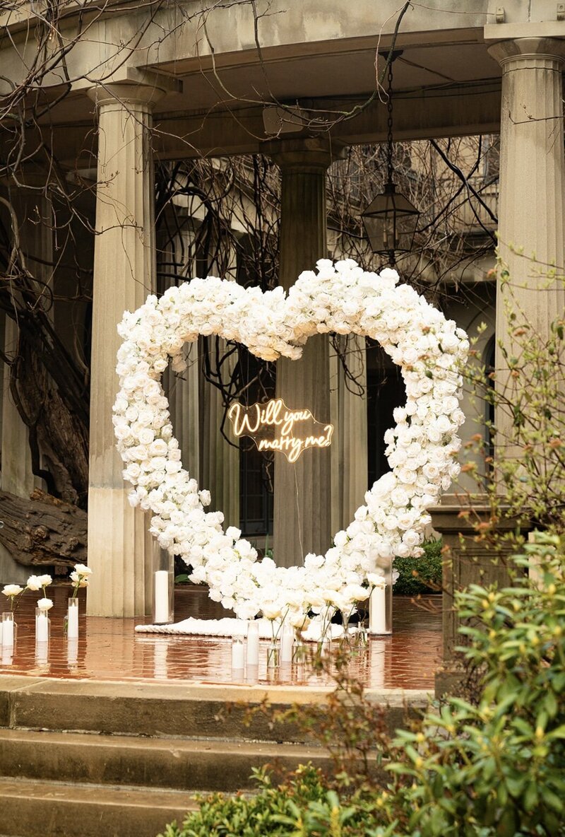 Romantic proposal backdrop with a large white floral heart arch and ‘Will you marry me?’ neon sign, styled by Yours Truly, a wedding planner and designer in New Jersey.
