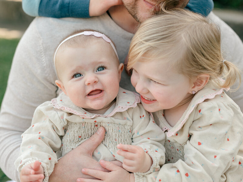 A heartwarming family moment featuring a father embracing his two young daughters. The baby, wearing a soft pink headband and a cream outfit with delicate details, gazes with bright eyes, while her older sister, dressed in a matching outfit, smiles joyfully beside her. Their father’s arms wrap around them, creating a warm and intimate connection. Captured in soft, natural light, this candid family portrait embodies the storytelling essence of KCP Families' photography