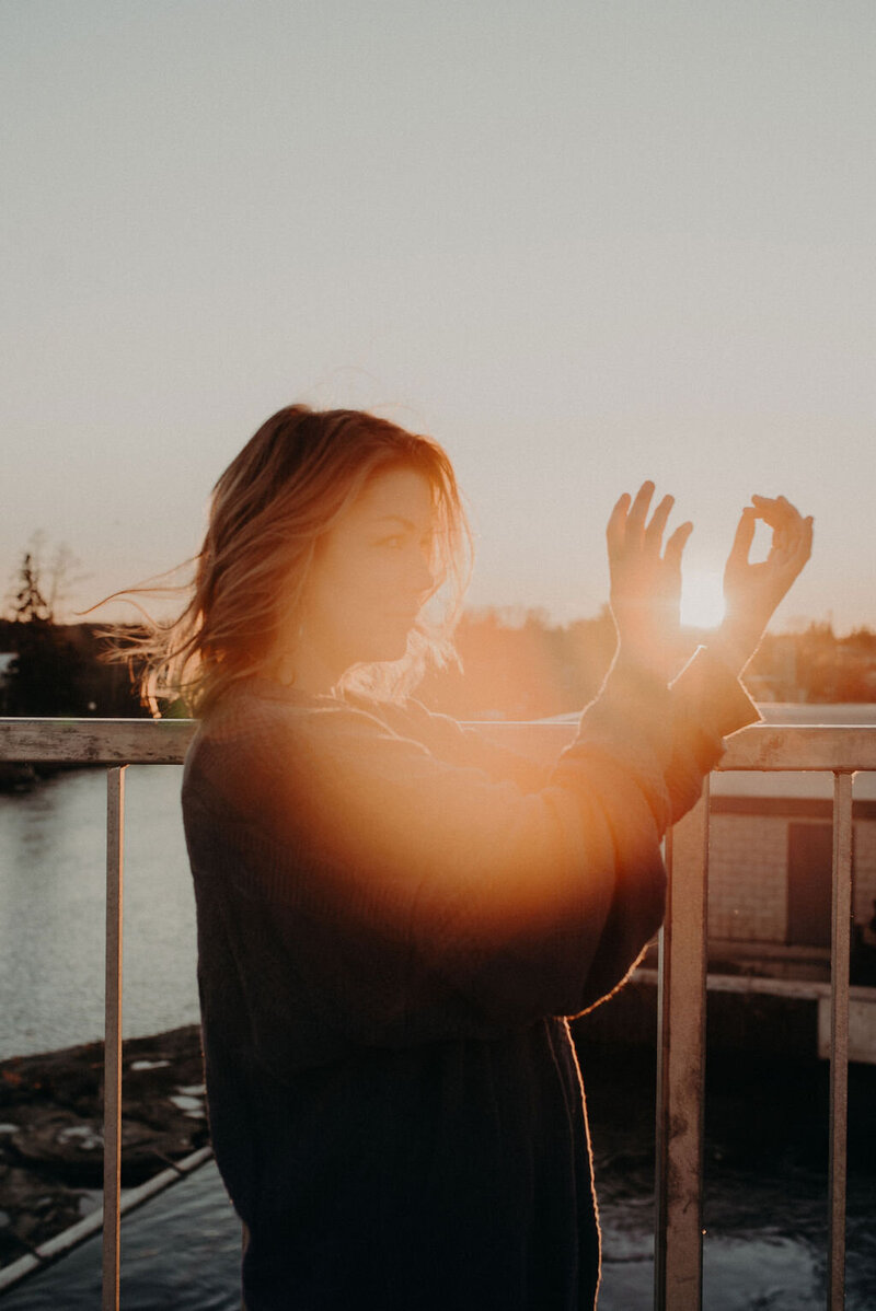 A woman holds her hands up in front of a setting sun.