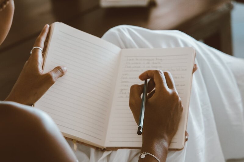 A woman in white pants writing in her journal with a pen in her hand.