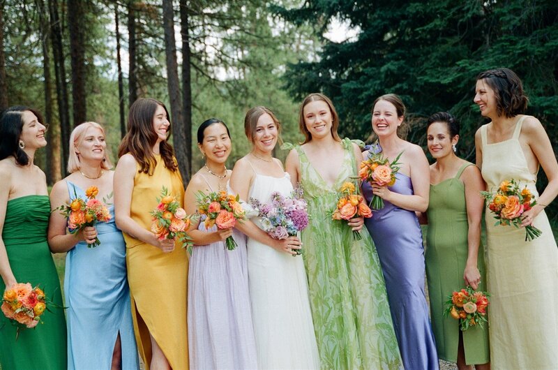 Bride Torrie with bridesmaids holding contrasting bouquets—lavender and blue for the bride, bold orange mono-floral bouquets for the bridesmaids—at a forest wedding, by Summer Robbins Flowers in Central Oregon