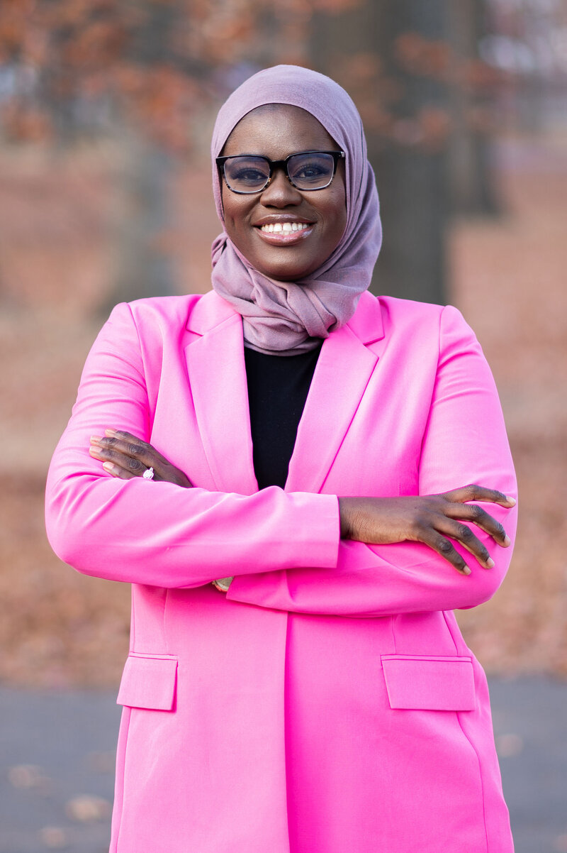 Photo of Isatou Bittaye Job, a grant writer, dressed in a pink blazer with a black shirt underneath , standing in an outdoor setting. She is smiling widely, and wearing black-framed glasses and a purple headwrap.