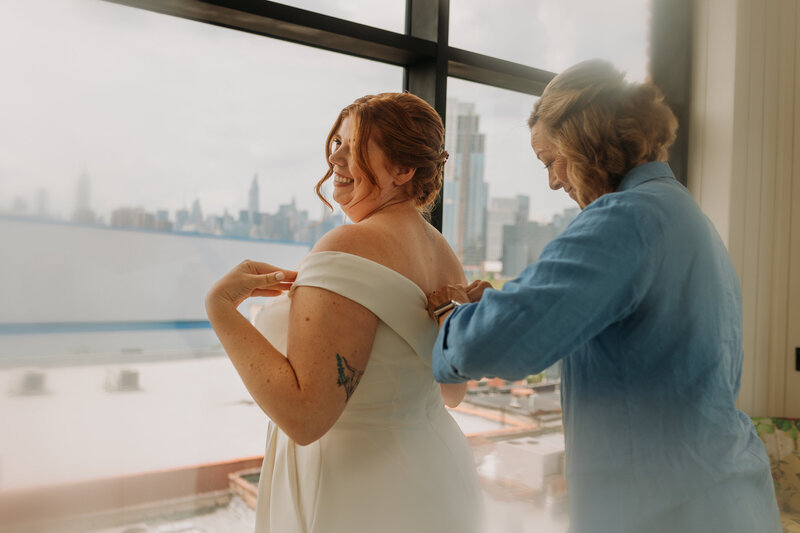 Bride smiles as her mother helps her into her bridal suit overlooking Manhattan skyline in Williamsburg Hotel room