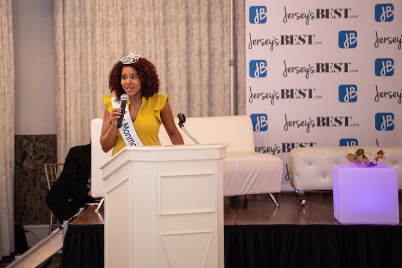 Miss Monmouth in a mustard dress with her sash and crown speaking at a podium as a keynote speaker at an International Women's Day Business Summit event hosted by NJ Media and the North Jersey Chamber of Commerce.
