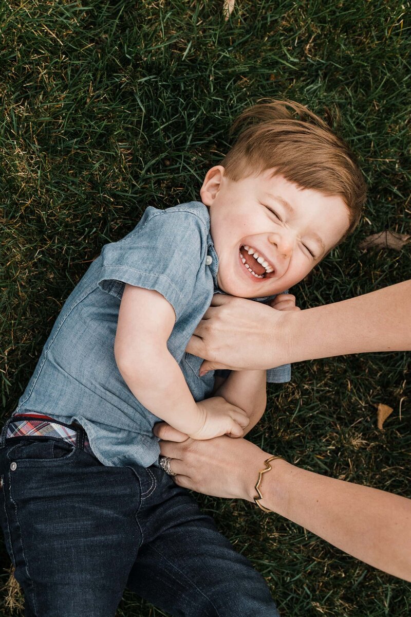 A young boy laughing joyfully while lying on grass during a family photography session.