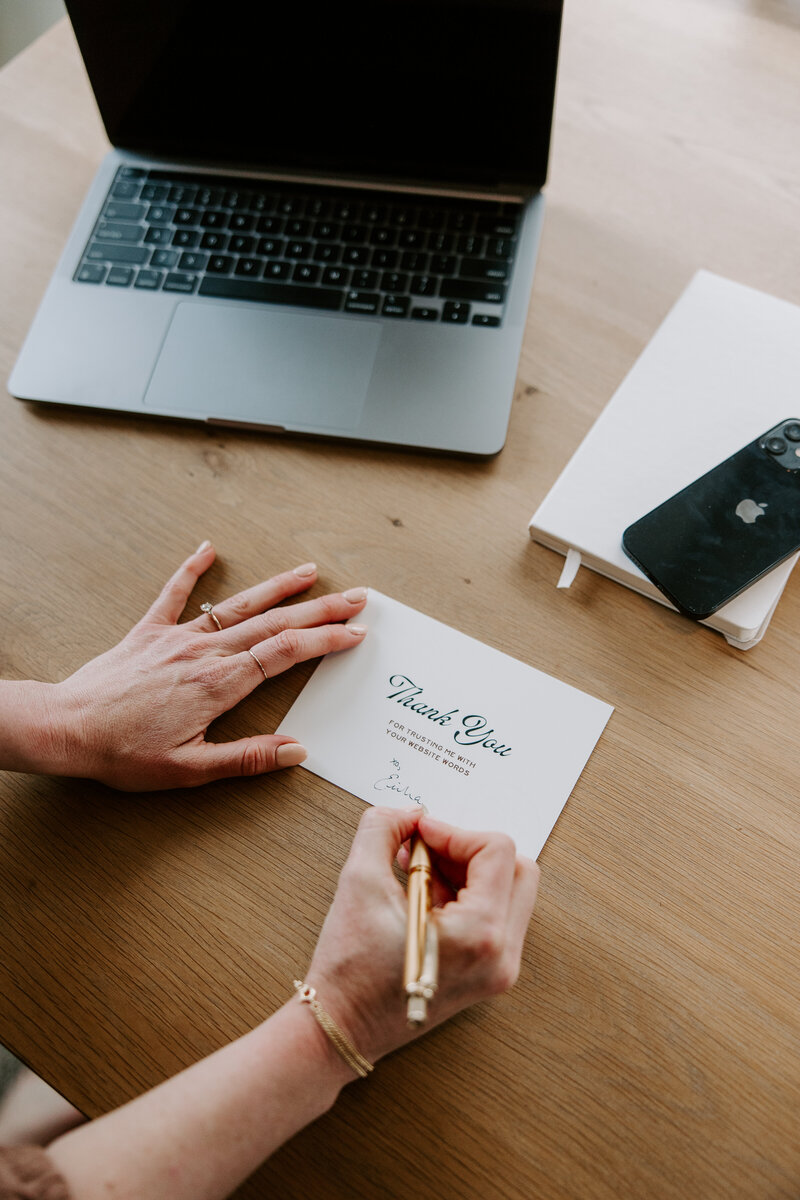 Website copywriter Erika Fitzgerald writing a thank you note at her desk with a laptop, showcasing her Showit website