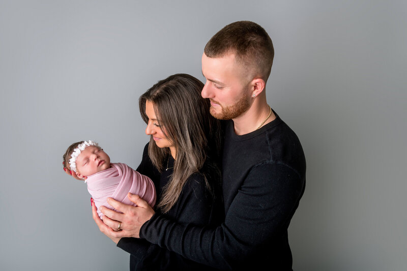 Connecticut newborn photographer captures tender studio portrait of new parents in black holding sleeping baby girl wrapped in pink with floral headband