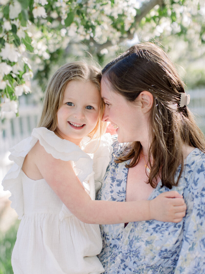 Closeup of a mom holding her daughter and nuzzling in close while girl is smiling, in a spring garden under flowering cherry trees by NH family photographer Fieldstone Studio.
