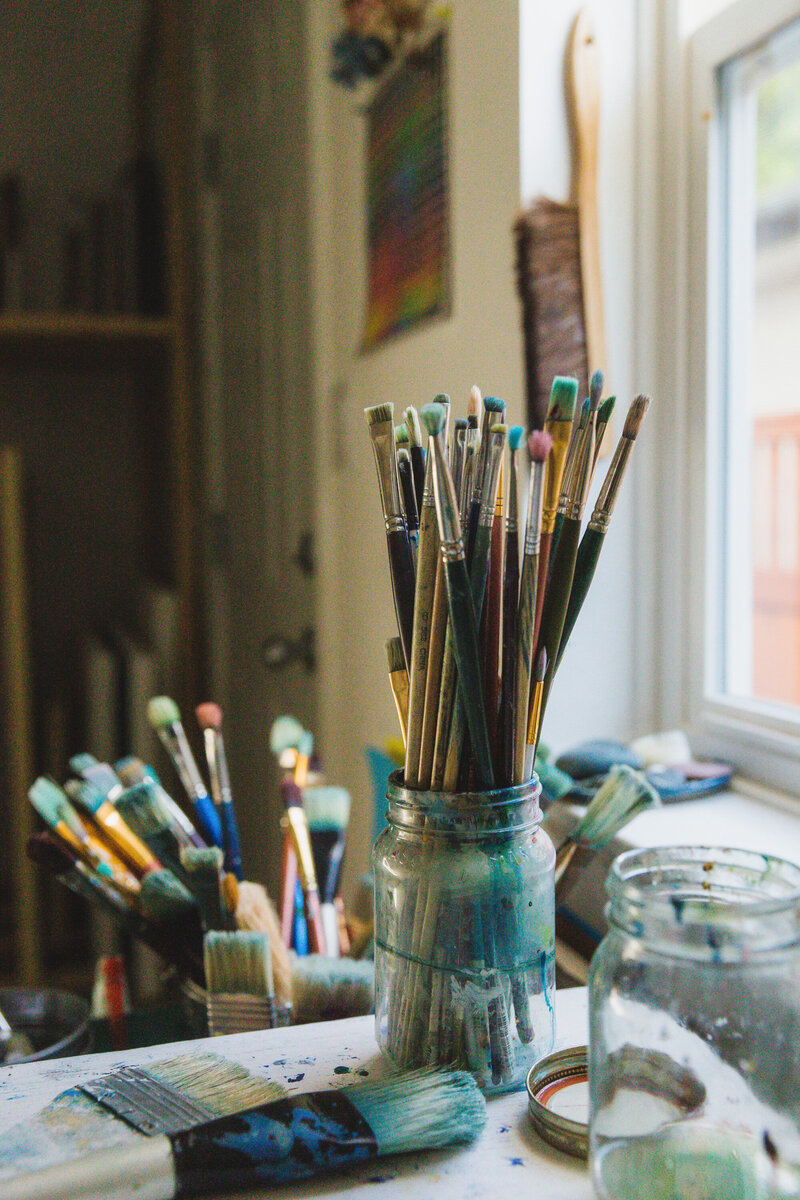 A close-up of a jar filled with well-used paintbrushes, their bristles stained with shades of blue and green, sitting on a paint-splattered table in an artist’s studio. Additional brushes and art supplies are scattered in the softly lit background near a window, creating a cozy and creative atmosphere.