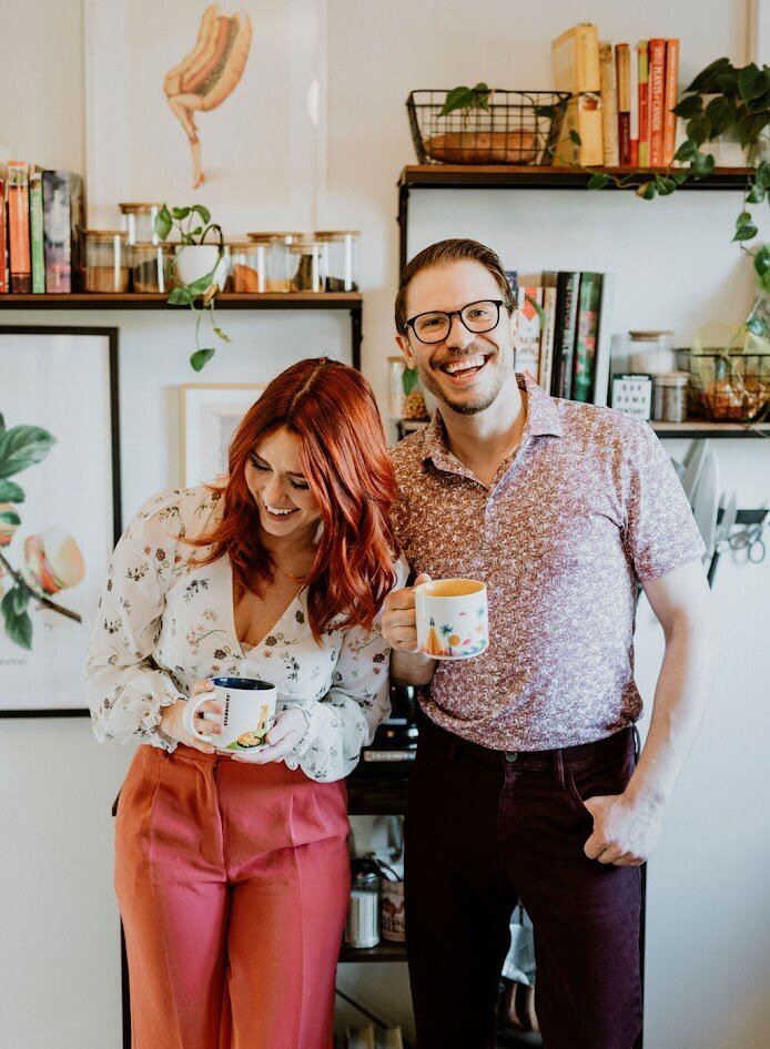 Brittany Frid and Lee Large, wedding and event designers and decorators in Ottawa and Prince Edward County, are standing in their studio kitchen smiling and laughing over coffee.