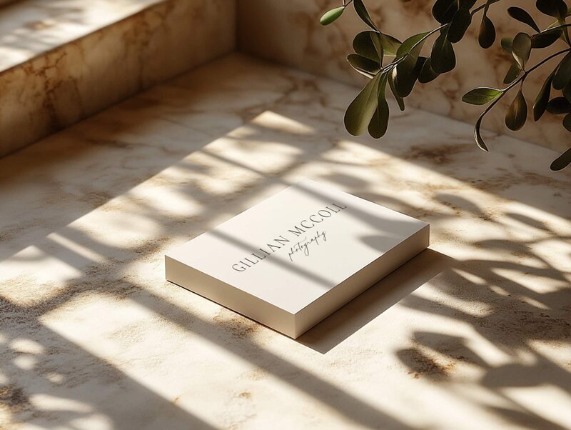 A closed book with "Gillian McColl" written on the cover is placed on a marble surface, embodying the elegance akin to showit web design for luxury photographers. Natural light and shadows from nearby plants fall across the book and surface, adding a touch of brand and identity services flair.