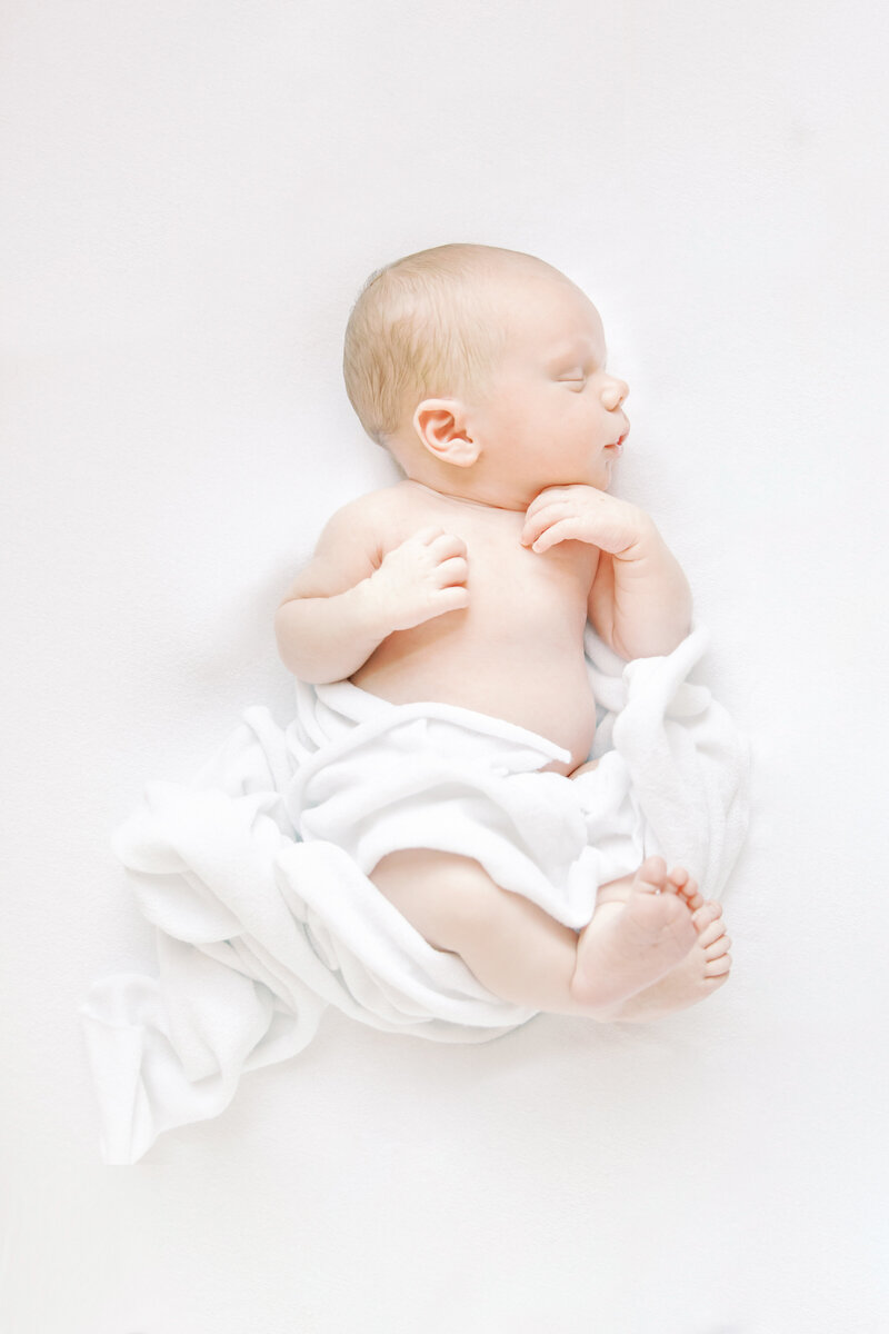 Baby in a neutral onesie sitting up on a white bed in a bright studio setting.