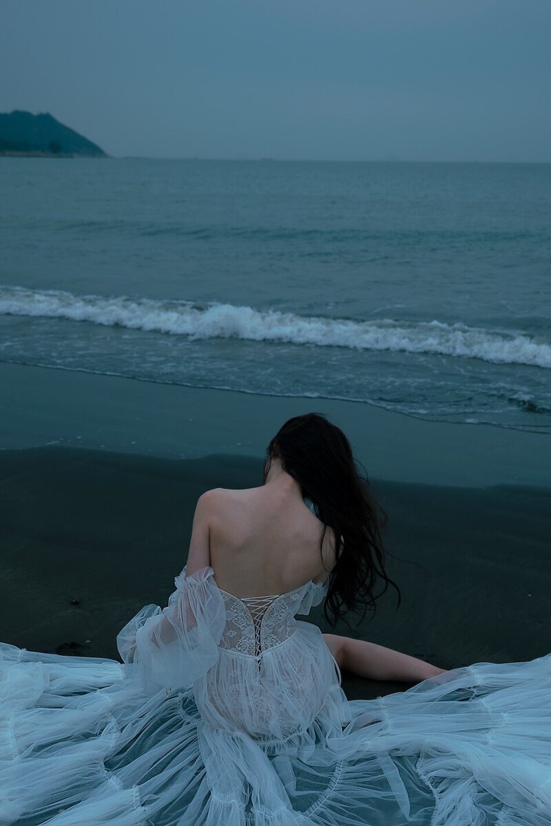 A woman wearing a delicate sheer couture dress shot on the beach in dark moody tones.