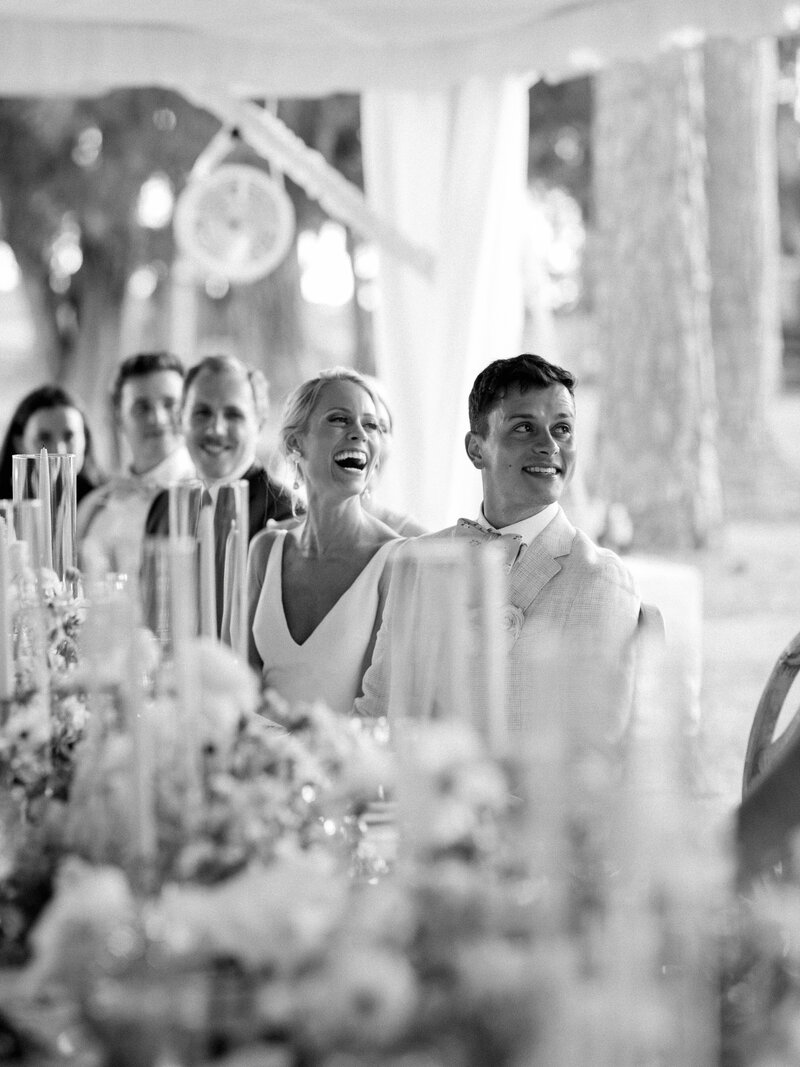 A black and white photo bride and groom seated at their reception in Charleston, SC laughing at a guest out of frame during their toast.
