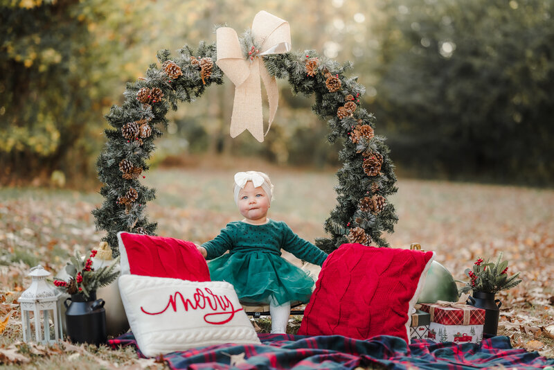 A toddler wearing a dark green dress sits on a large Christmas wreath. She is surrounded by Christmas decorations during her Christmas Session in Chesapeake.