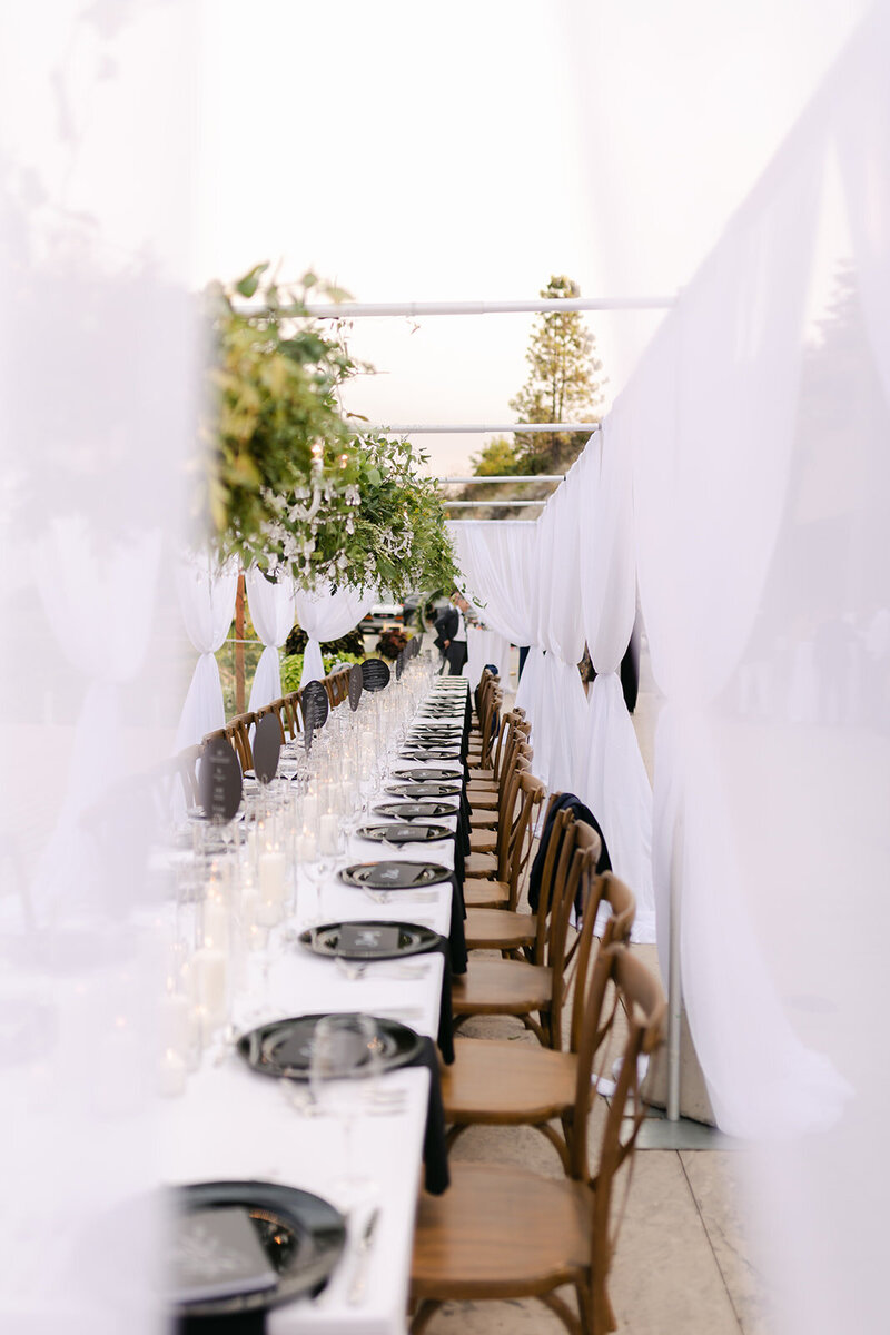 A long table wedding design includes white drapes, greenery, crystal chandeliers, black plates and white candles.