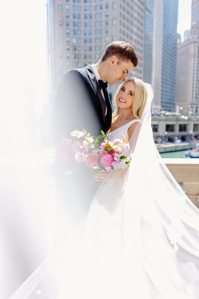 Bride and groom embrace as veil flows in the breeze and the Chicago skyline behind them