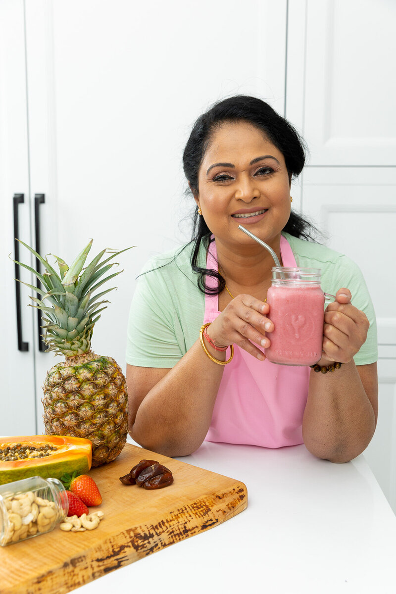 Kalinie holding a pink smoothie with a veriety of fruit on the counter next to her.