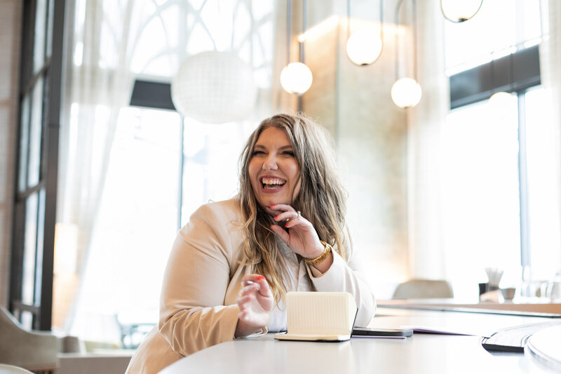 Ashleigh Henry Life Coach and Speaker at Kimpton Hotels Arras at the Bar Area, writing in a small notebook while smiling at the camera, wearing a tan blazer and loose curls with gold jewelry.