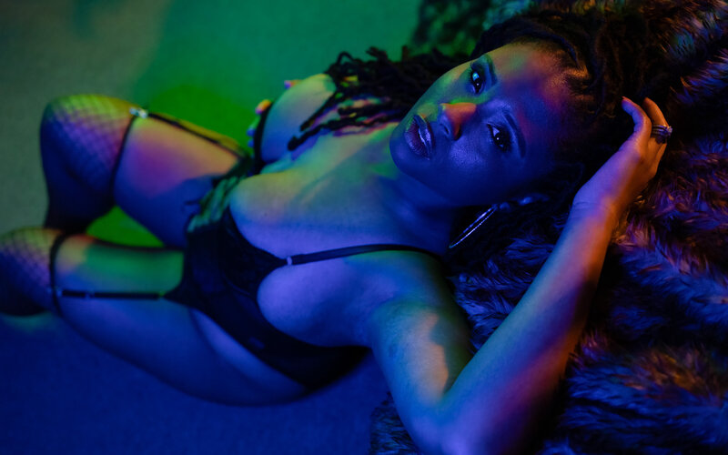 A woman in a colorfully lit room sits on the floor in lingerie with a hand in her braids