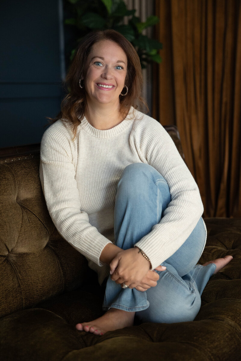 Christina Barnett, Monroe Georgia photographer, smiling and relaxed in a personal branding photo, wearing a cream sweater and light blue jeans, sitting barefoot on a vintage brown velvet couch in a cozy, warmly lit indoor studio setting.
