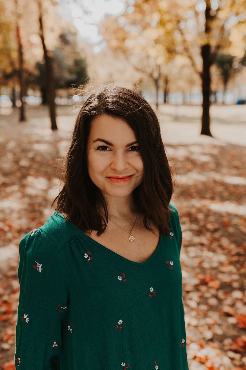 Sarah Berneche, brown-haired woman in a turquoise dress, stands in High Park with trees and autumn leaves in the background. She is a Toronto Intuitive Eating Counsellor specializing in binge eating, emotional eating, and body image.