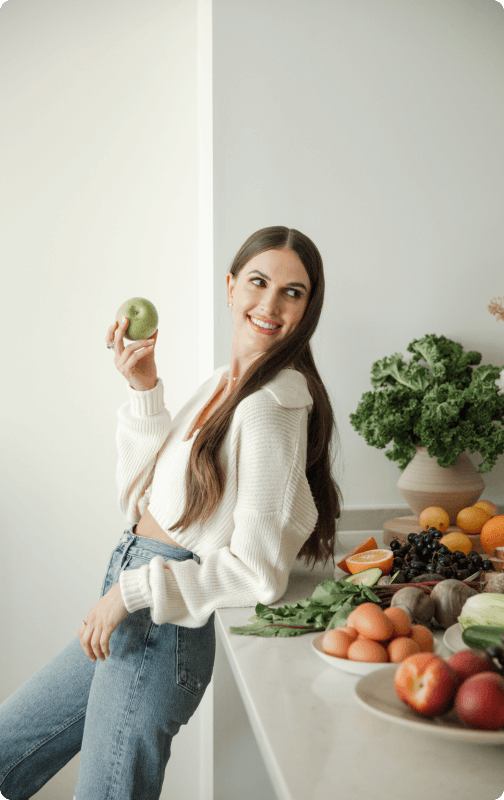 Anya Rosen, founder of Birchwell Clinic, holding a green apple and leaning a against a kitchen  countertop that has a display of leafy greens, colorful fruits, and eggs.
