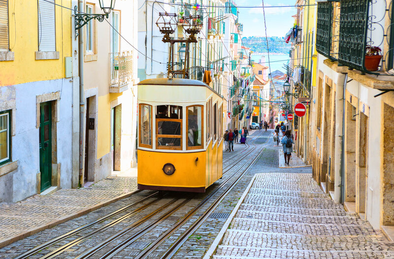 Historic yellow tram traveling up a steep cobblestone street lined with colorful buildings and pedestrians, with a view of the water in the distance.