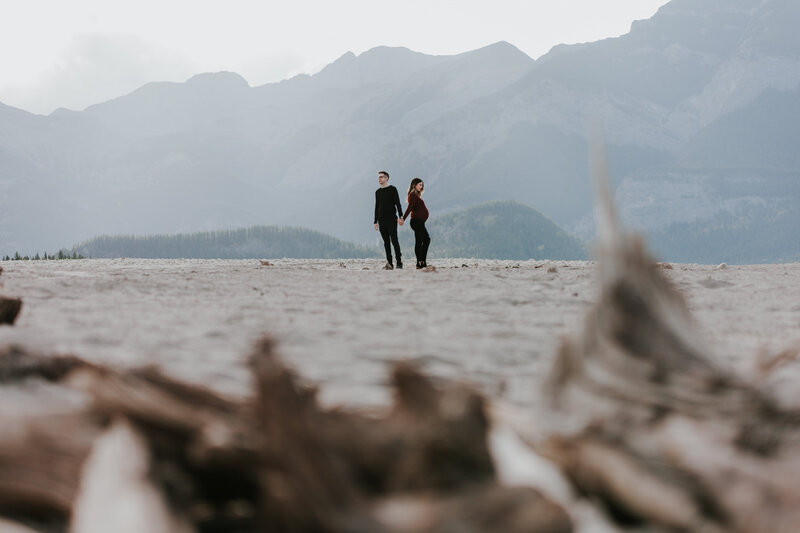 A long distance shot of a man and pregnant wife standing, holding hands, in front of a mountain backdrop