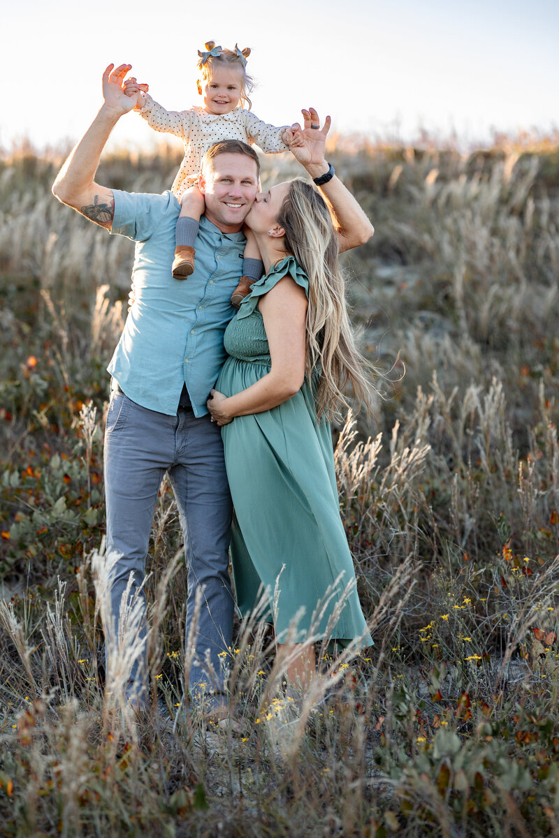 A joyful little girl sits on her father's shoulders, while her parents share a warm embrace below.  Captured in a sun-drenched field in Virginia Beach by Brooke Tucker Photography, this playful family portrait radiates love, laughter, and golden hour magic. 