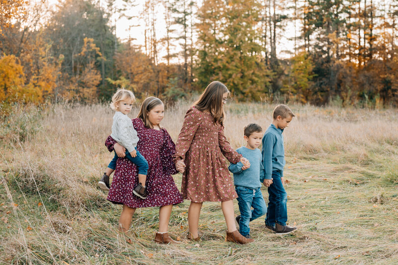 5 siblings holding hands while walking in a field surrounded by fall colors.