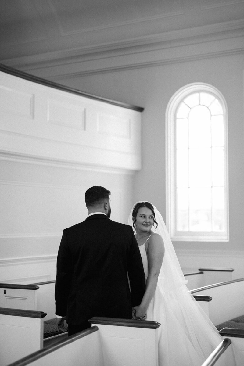 Bride looking back at the groom while walking inside Martha-Mary Chapel with pews and arched windows in view.