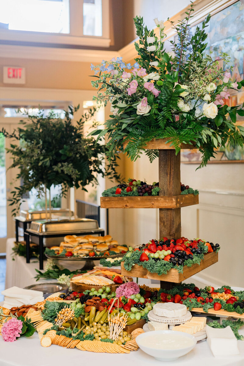 A buffet table with a wooden tiered stand holding assorted fresh fruits and leafy floral arrangements, surrounded by cheese, crackers, vegetables, and a tray of sandwiches in a bright, elegant room.