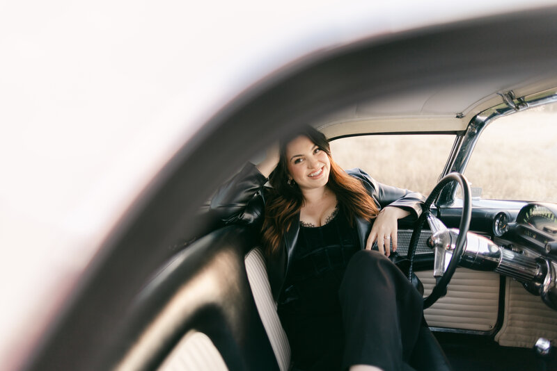 Woman smiling inside a retro car with her arm resting on the window frame, styled in a leather jacket and velvet top. Editorial portrait photography in Greenville, South Carolina by Dani Lacey Photographs.