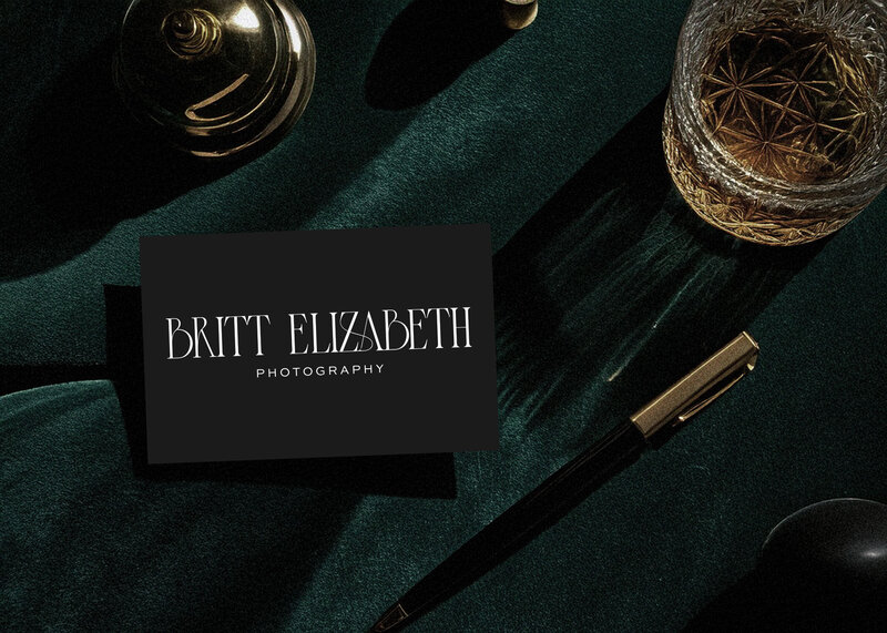 A business card reading "Britt Elizabeth Photography" rests on a dark green surface beside a gold pen, a glass of liquor, and a decorative brass object—reflecting refined brand and web design for small businesses.
