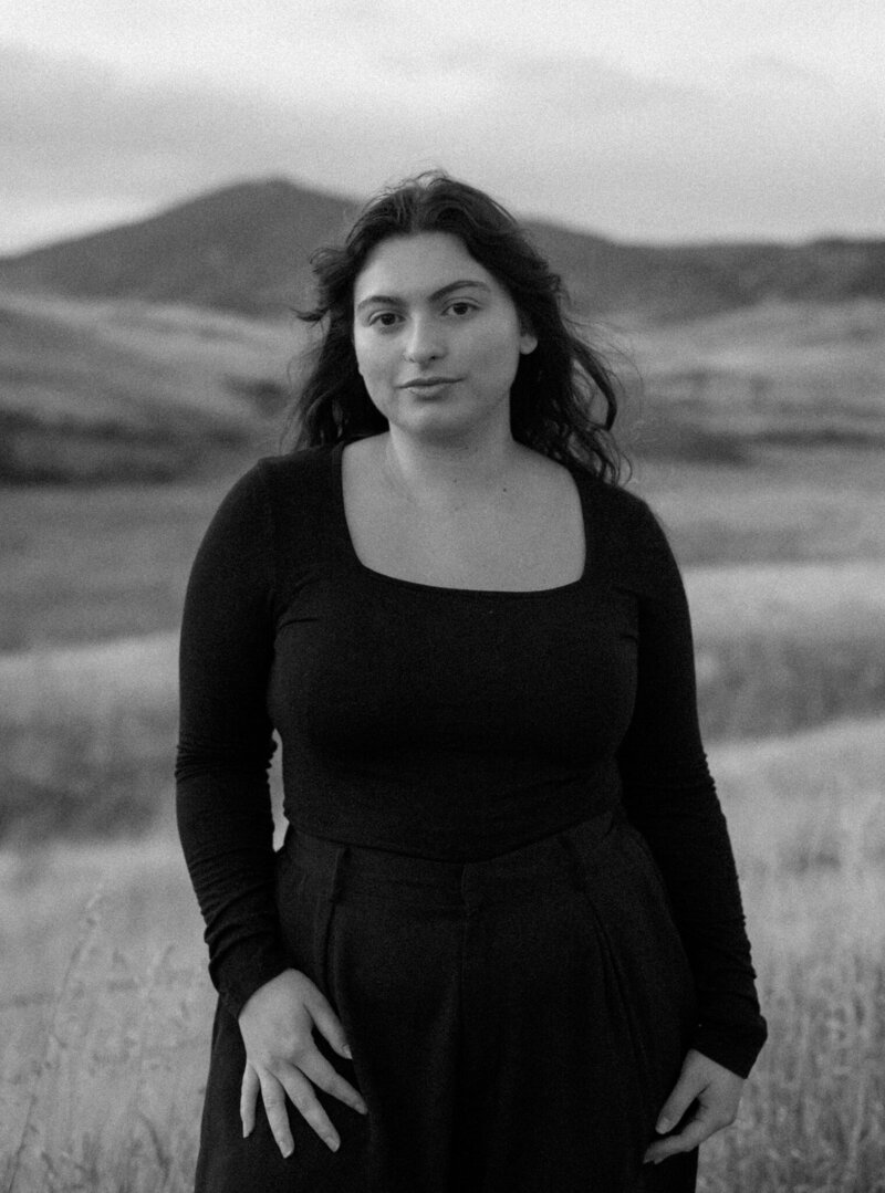 A black-and-white portrait of Daniella Diaz standing in an open field with rolling hills and distant mountains in the background. She wears a long-sleeved black top with a square neckline and high-waisted trousers, her dark wavy hair gently tousled by the wind. Her expression is calm and introspective, with a soft yet confident gaze directed at the camera. The grainy texture of the photograph adds a timeless, cinematic feel, while the blurred landscape behind her enhances the sense of depth and solitude. The natural setting and monochrome tones create a moody, artistic portrait that evokes a quiet strength and connection to nature.