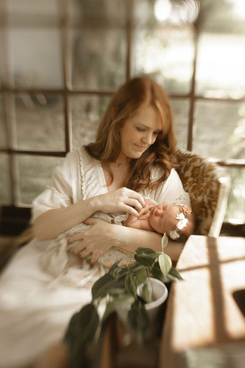 A mother sitting in a vintage chair holding her newborn baby girl and admiring her sweet details. 