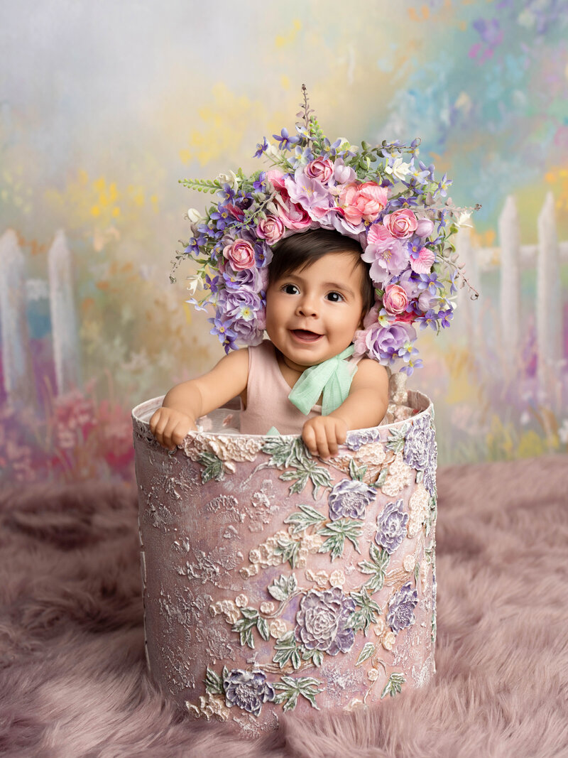 A baby with a serene expression wears a vibrant flower crown of roses in purple, pink, and yellow. The soft pink blanket adds a cozy, gentle backdrop.