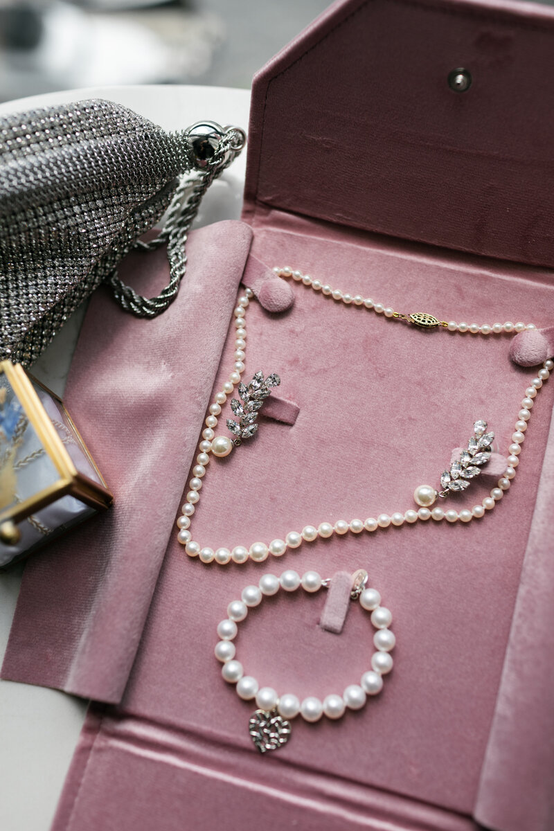 flat lay of bride's pearl jewelry in a blush velvet jewelry holder nest to a glass ring box and silver sparkly clutch purse