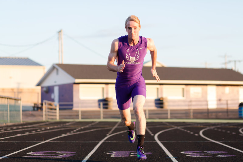 triway senior guy in track uniform running towards camera full speed, photographer is sitting on ground so he looks much larger than life, photographed at Triway High School, photographed by Jamie Lynette Photography Canton Ohio Senior Photographer