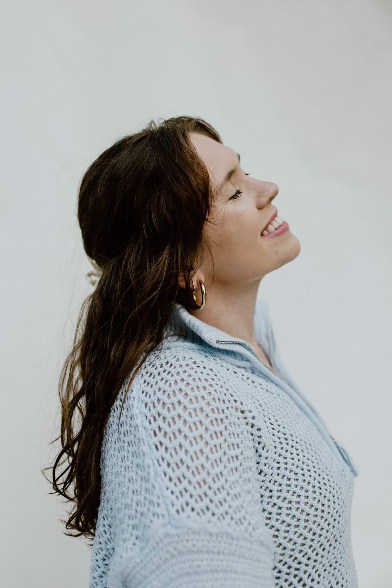 A girl turned to the side smiling up at the sky with her eyes closed. Wearing gold hoops and a light blue sweater.