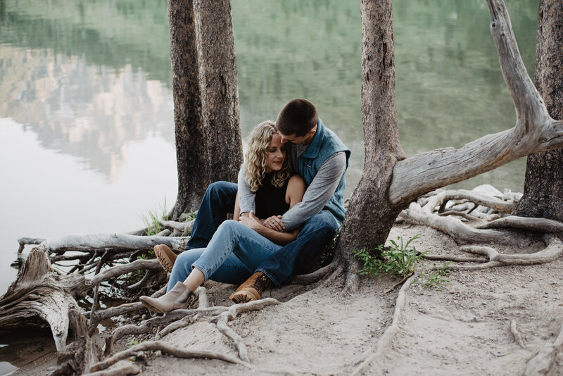 jackson hole photographers captures engagemtn pictures with couple sitting together on exposed roots of a tree on a lake