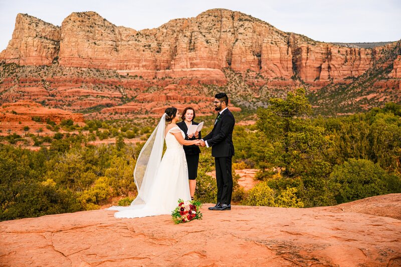 Bride and groom dressed in wedding attire during holding hands looking at each other during elopement ceremony on red rocks Bell Rock in Sedona Arizona