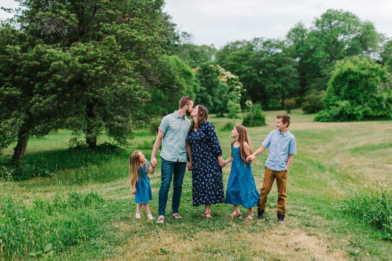 Family of five holding hands in a field while mom and dad kiss each other